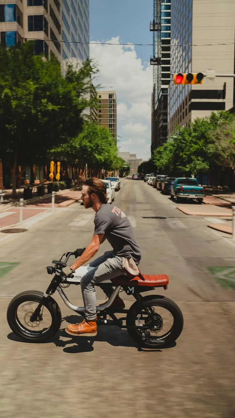 A lineup of Monday Motorbikes electric motorcycles parked in a modern urban environment. Café-racer inspired silhouettes with exposed frames, premium finishes, and minimalist details. Riders in stylish urban gear, city architecture in the background.
